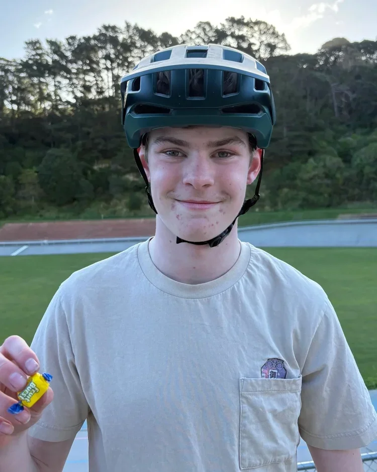 A young man wearing a bike helmet and casual t-shirt, smiling outdoors on a sports field with a background of trees and sky, holding a small candy in his hand.