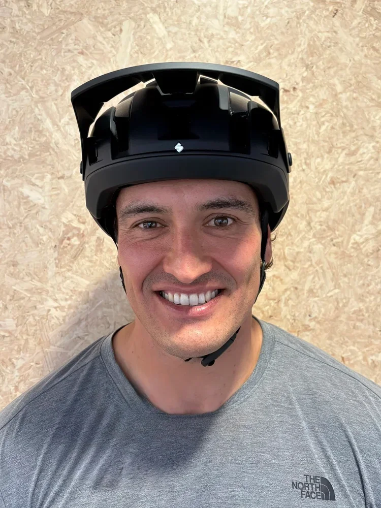 A smiling man wearing a black helmet and a gray The North Face t-shirt standing against a beige wall.