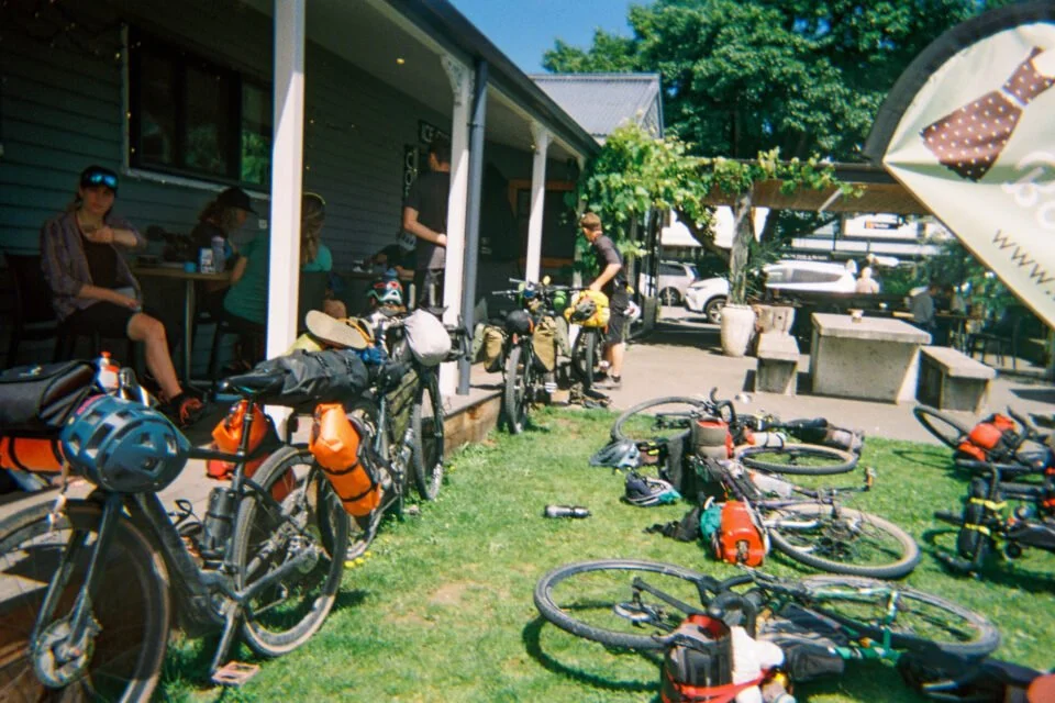 Bicycles with gear and helmets parked outside a cafe on a grassy area, with people sitting at outdoor tables and preparing for a bike ride.