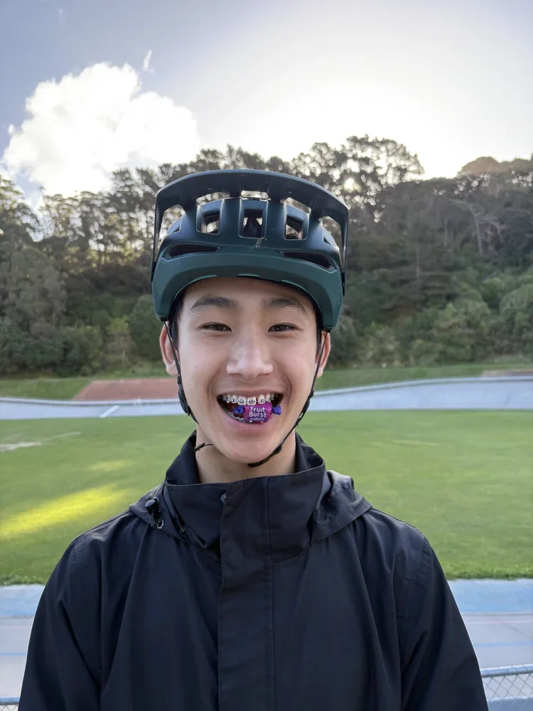 A young person wearing a helmet and black jacket standing outdoors at a track and field stadium with trees and a cloudy sky in the background, smiling with a colorful candy in their mouth.