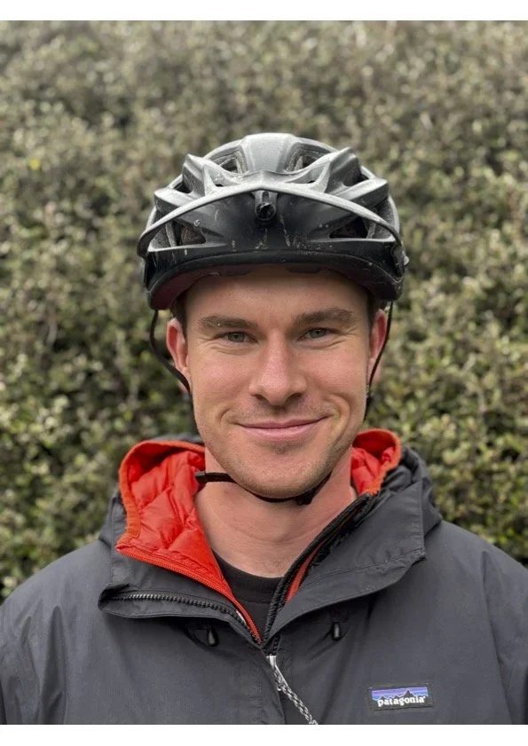 A young man smiling, wearing a black bicycle helmet and a black and red outdoor jacket, standing outdoors with a background of green bushes.