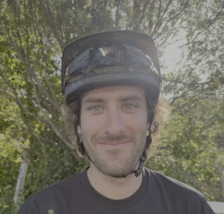 Young man wearing a black bicycle helmet and black shirt, smiling outdoors with green trees and sunlight in the background.