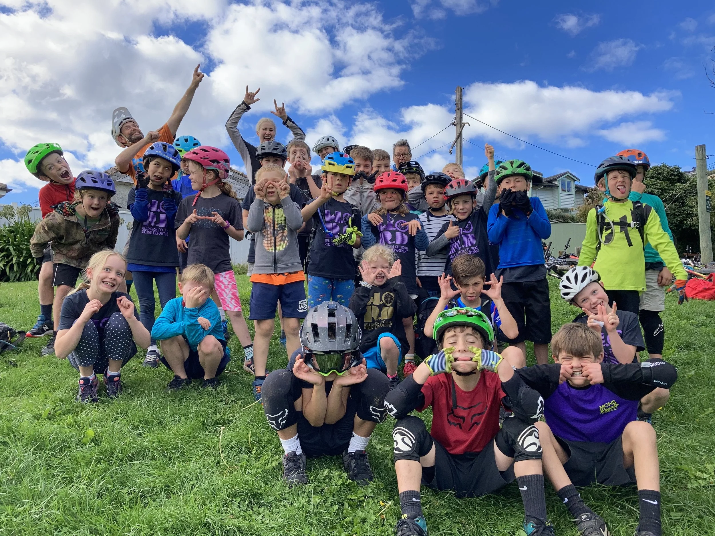A large group of children and a few adults, wearing helmets and sports gear, posing cheerfully outdoors on a grassy area under a partly cloudy sky.