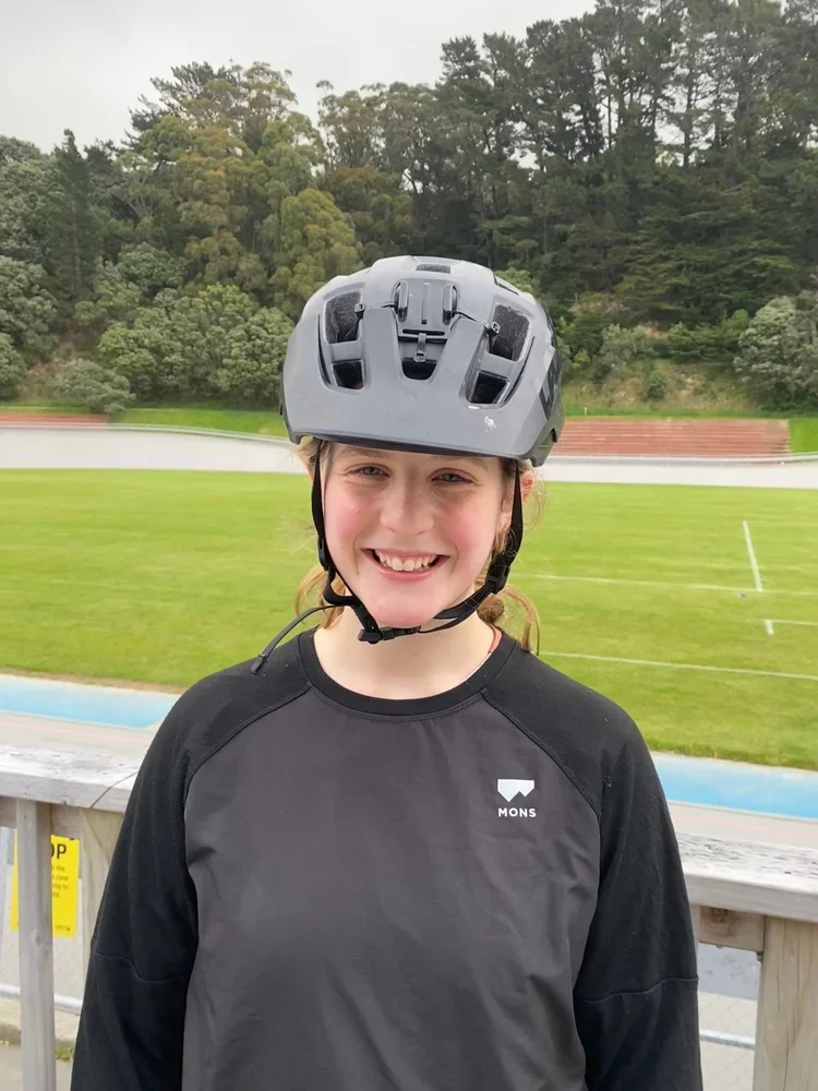 Young girl wearing a black and gray sports shirt and a helmet, smiling outdoors near a running track and grassy area with trees in the background.