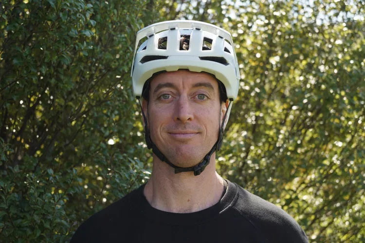 A man wearing a white bicycle helmet outdoors with green foliage in the background.