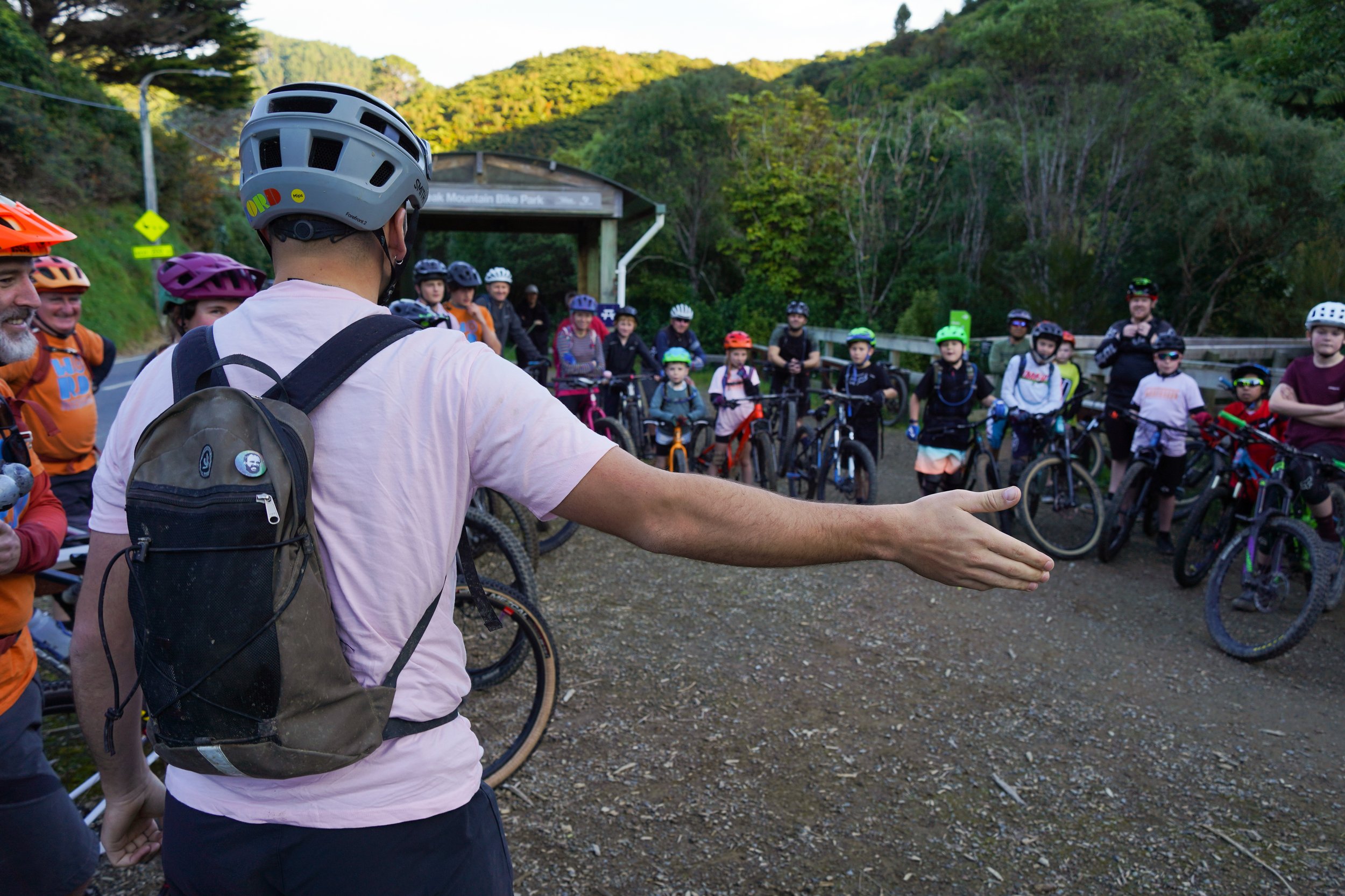 A group of children with adult supervisors gathered with their bicycles at the start or end of a biking event at MTB park surrounded by green trees in the mountains, as the adult in the foreground speaks to the group.