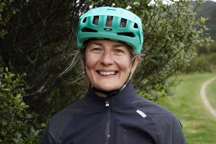 A smiling woman wearing a teal bike helmet and a black jacket outdoors with greenery and a trail in the background.