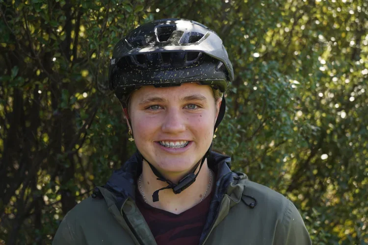 A smiling young man wearing a black bicycle helmet and a dark jacket outdoors with green foliage in the background.