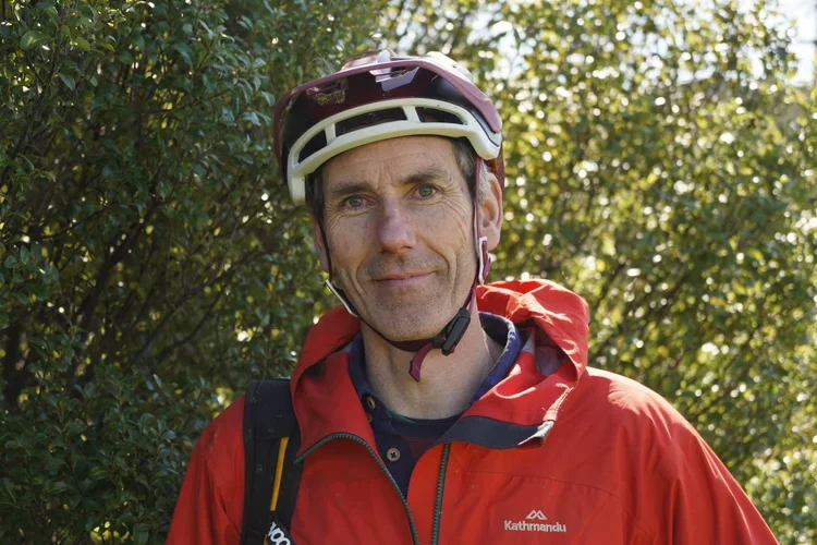 A man wearing a helmet and a red outdoor jacket stands outdoors with greenery in the background.