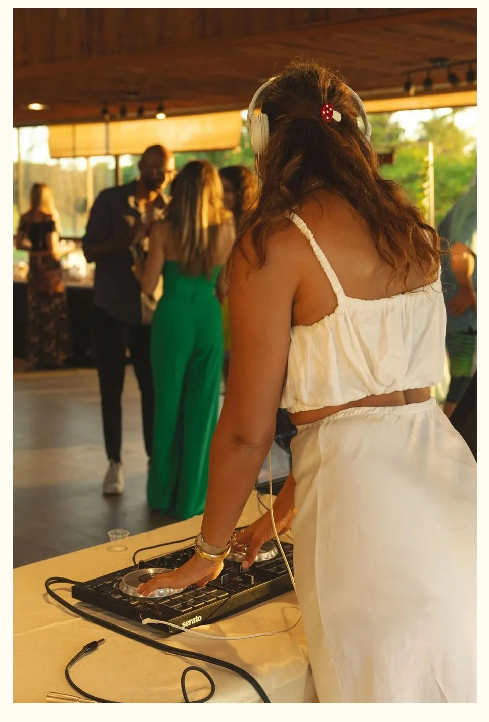 A woman wearing a white dress and headphones DJing at a party or event, with people dancing and socializing in the background.