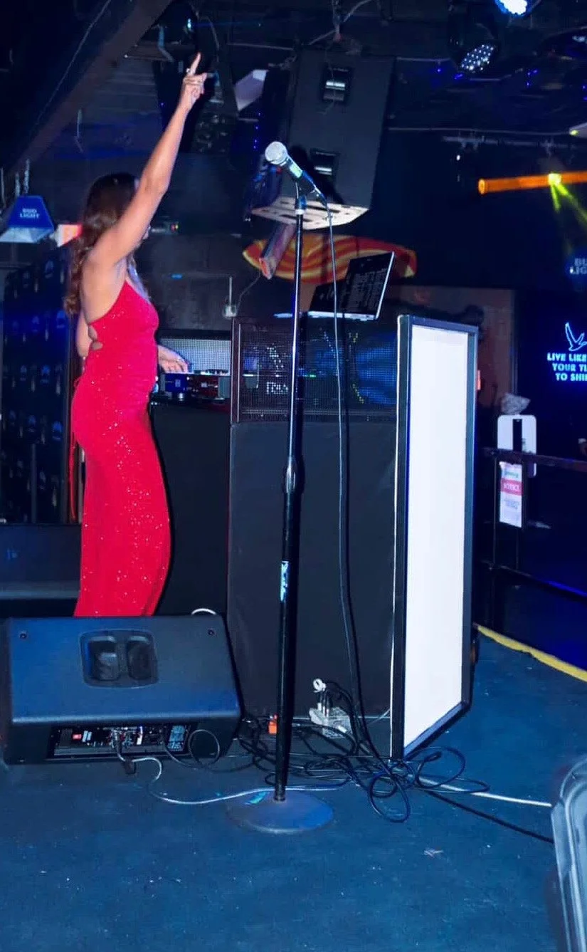 A woman in a red dress performing on stage with DJ equipment, microphone, and a laptop in a nightclub.