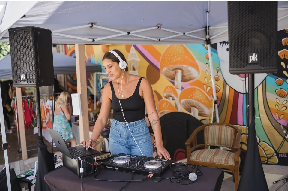 Female DJ wearing headphones, standing behind a DJ booth, mixing music with a laptop, at an outdoor event with colorful wall mural in the background.