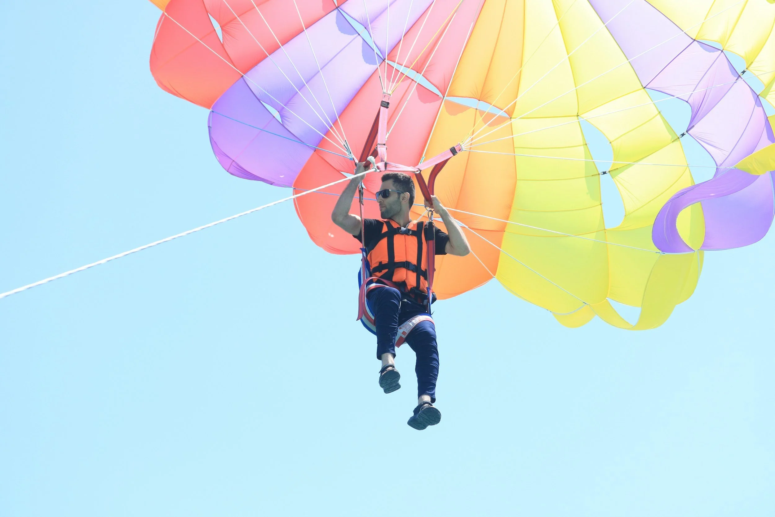 A man in sunglasses and a harness skydiving with a colorful parachute against a blue sky.