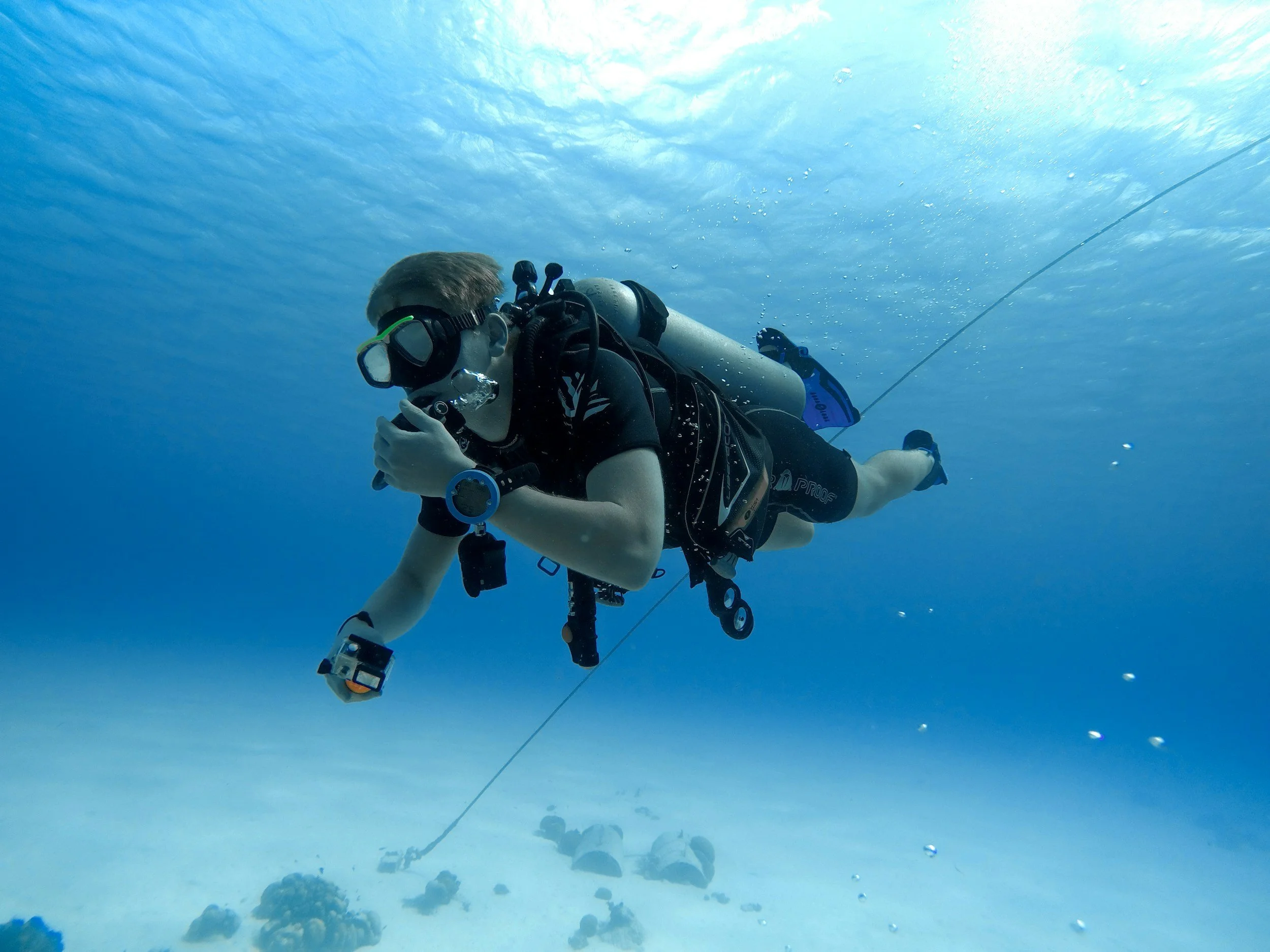 A scuba diver underwater holding a camera or device, wearing a mask and breathing apparatus, with a swimming line running diagonally in the background.