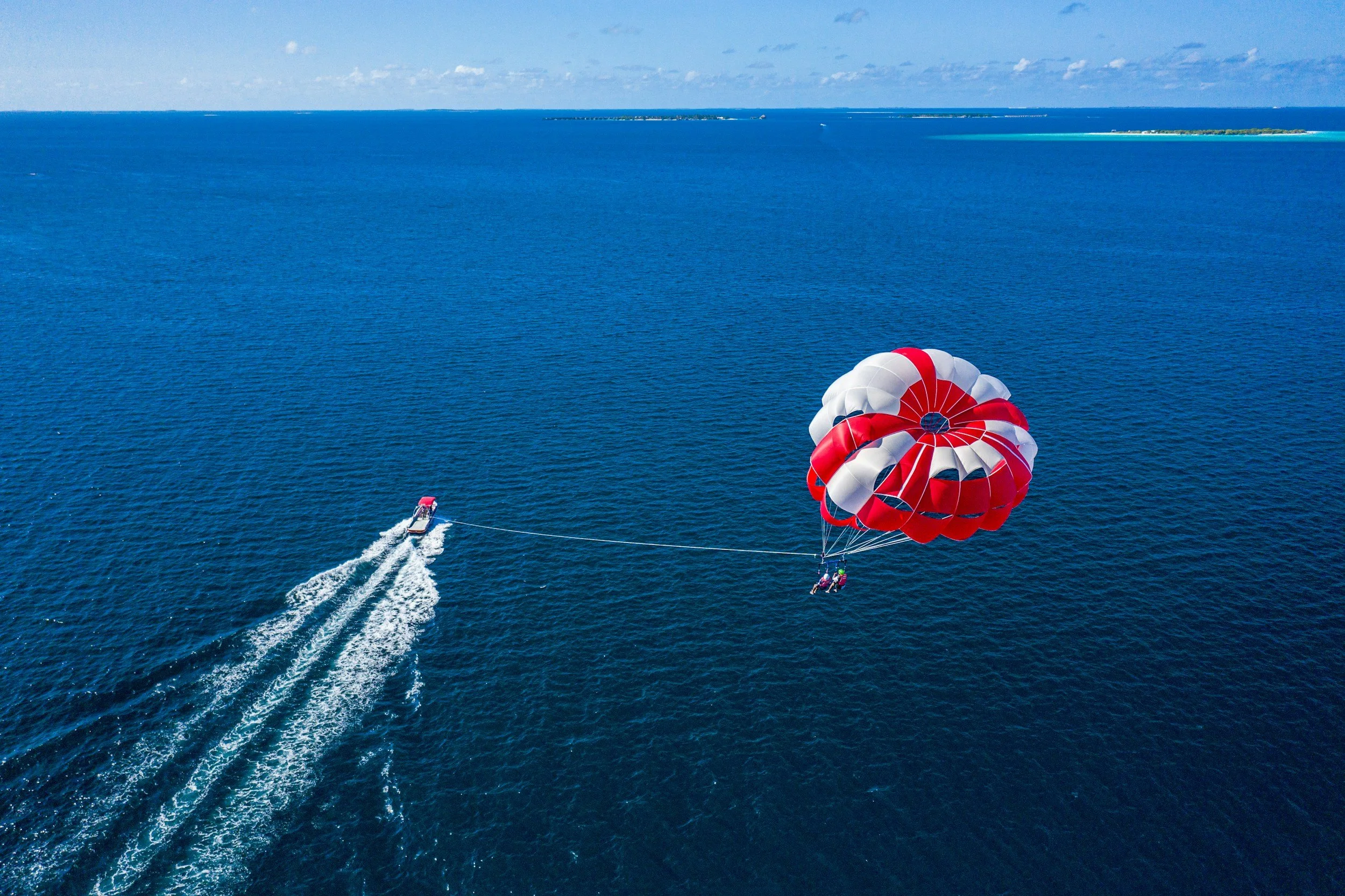 Aerial view of a parasailing adventure over a vast blue ocean with a boat towing a red, white, and blue parachute, and distant landmasses on the horizon.