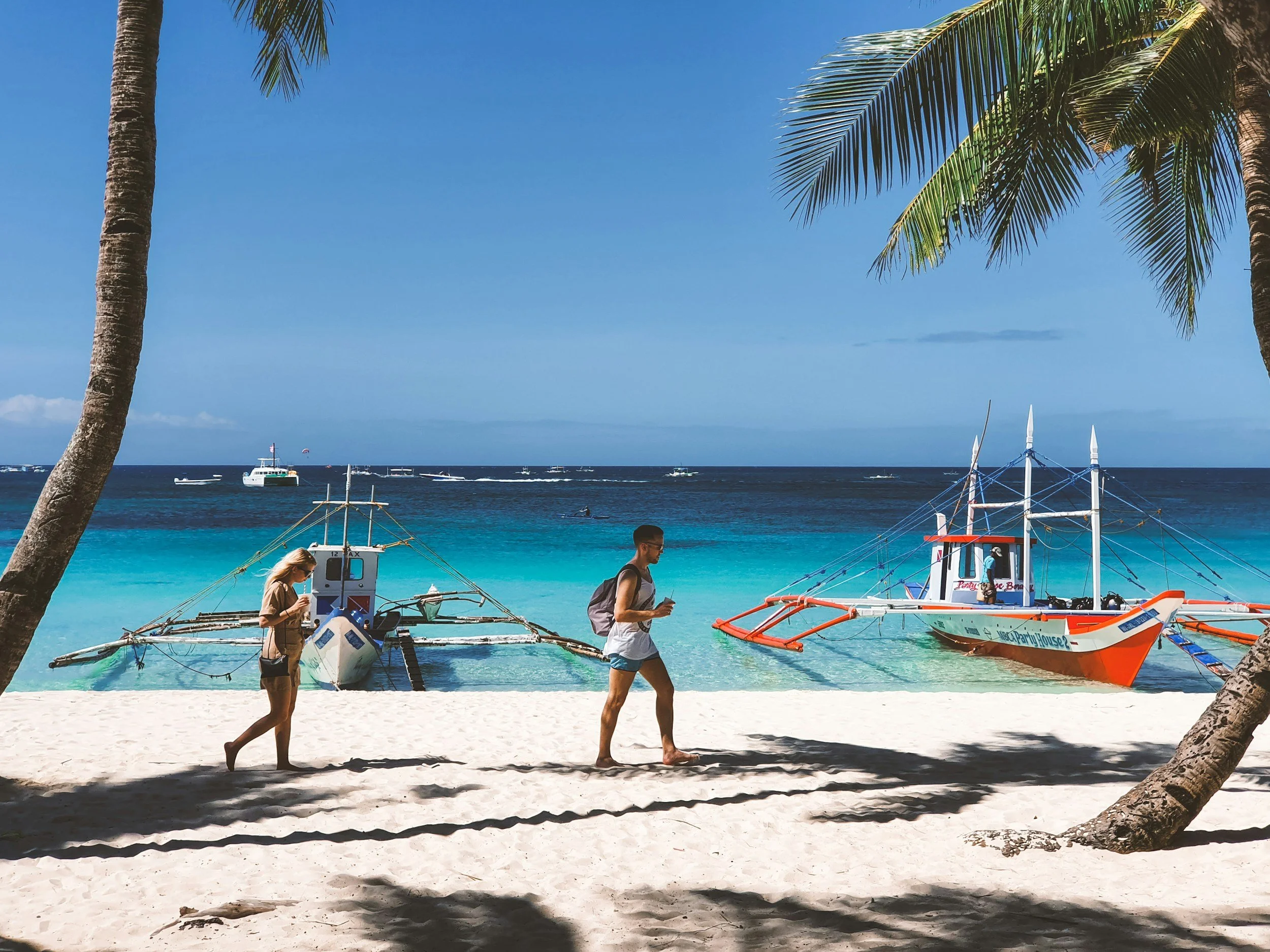 People walking on sandy beach near boats and clear turquoise water, with palm trees in the foreground and boats on the ocean, blue sky above.