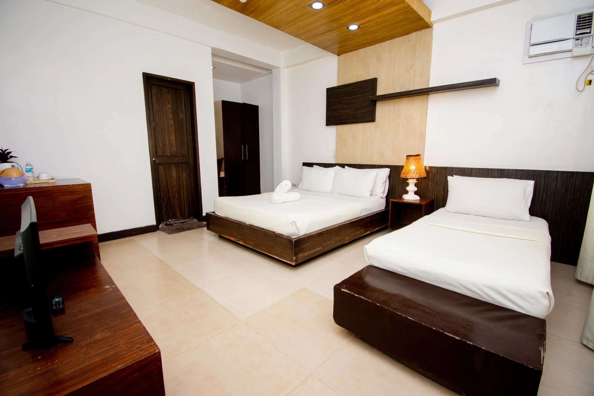 A bedroom with two beds, one with rolled towels and the other with pillows, dark wood furniture, a small nightstand with a lamp, a wardrobe, and an air conditioning unit on the wall.