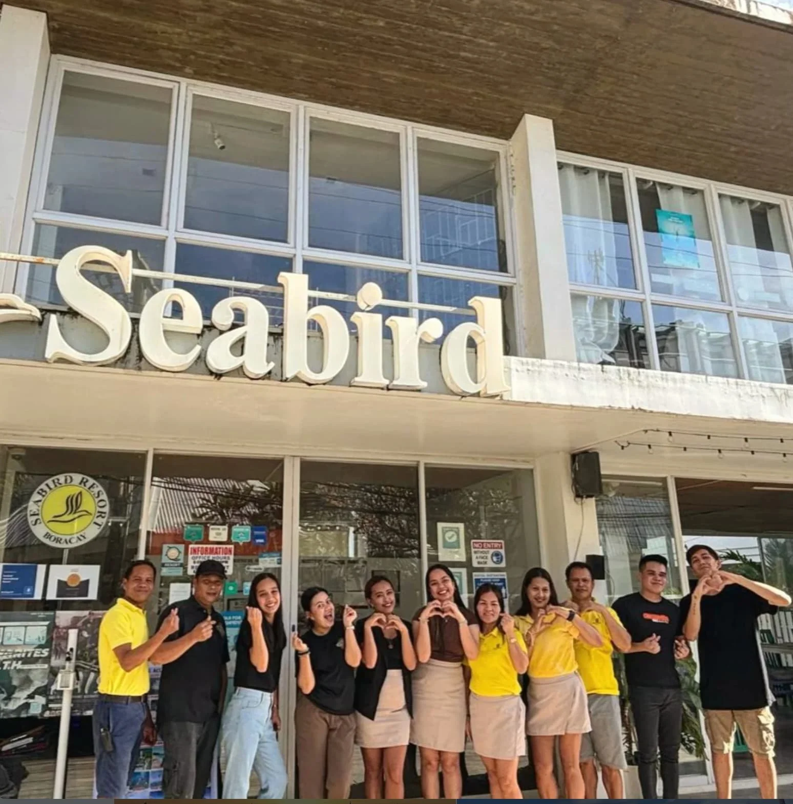 A group of people standing in front of a building with a sign that says 'Seabird' and some people making heart gestures with their hands.