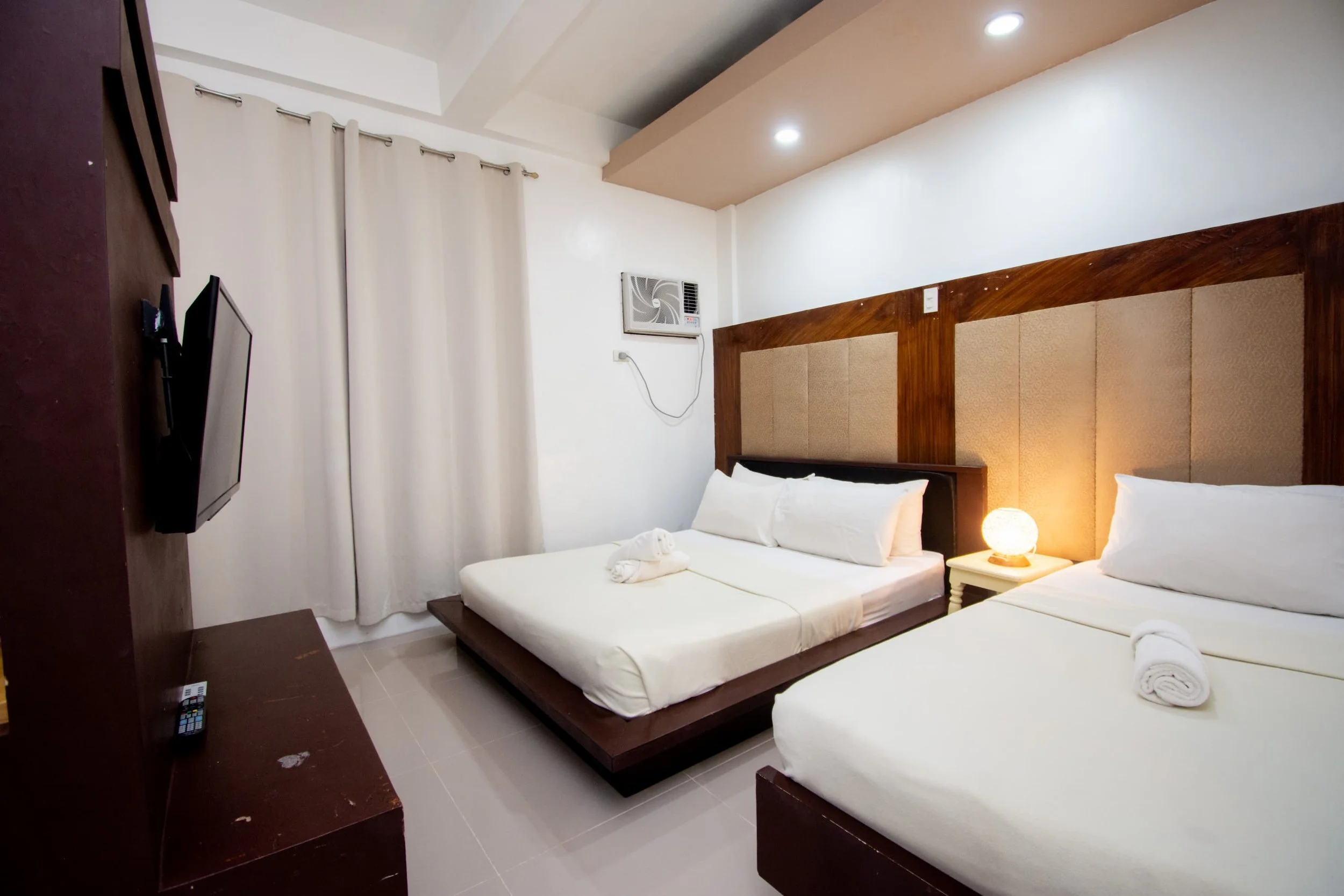 A hotel room with two beds, a headboard, a nightstand with a lamp, a wall-mounted TV, and an air conditioning unit on the wall.