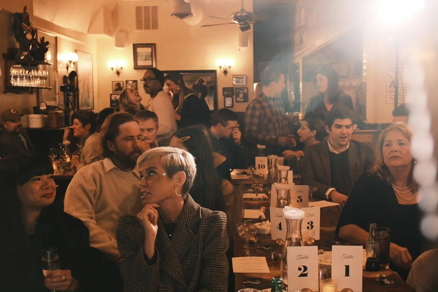 People gathered in a cozy restaurant, some seated at tables with table numbers, others standing and socializing, with warm lighting and vintage decor.