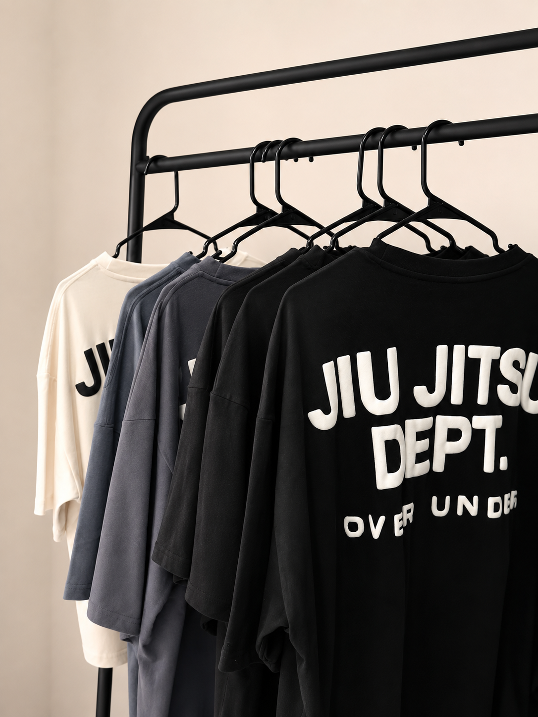 A clothing rack displaying four T-shirts in white, gray, dark gray, and black, with the black shirt featuring bold white text that reads 'JIU JITSU DEPT. OVER UN DE'.