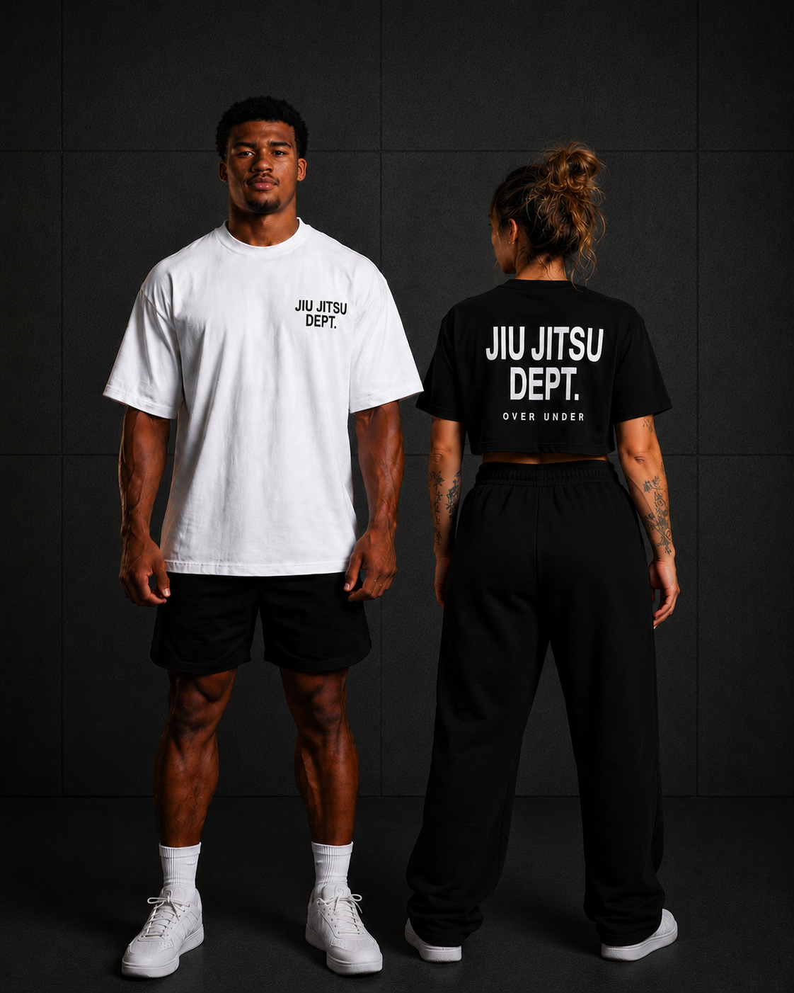 Two people standing against a black wall, facing away from the camera. The person on the left is a man wearing a white T-shirt with 'JIU JITSU DEPT.' written on it, black shorts, white socks, and white sneakers. The person on the right is a woman in a black cropped T-shirt with the same text, black sweatpants, and white sneakers, with her hair in a messy bun and visible tattoos on her arms.