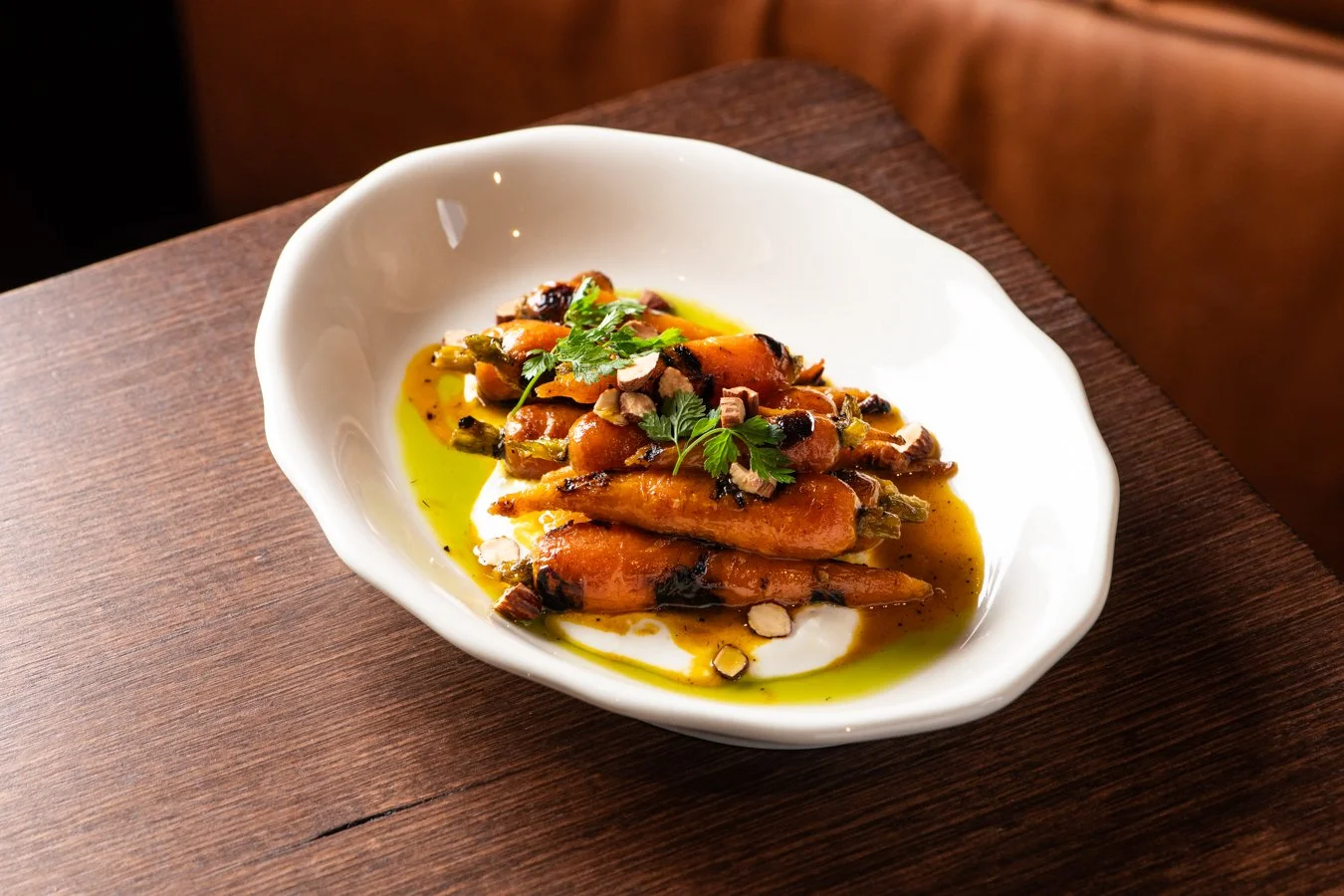 A white bowl of roasted carrots garnished with herbs and nuts, served with a green sauce drizzled on top, placed on a wooden table.