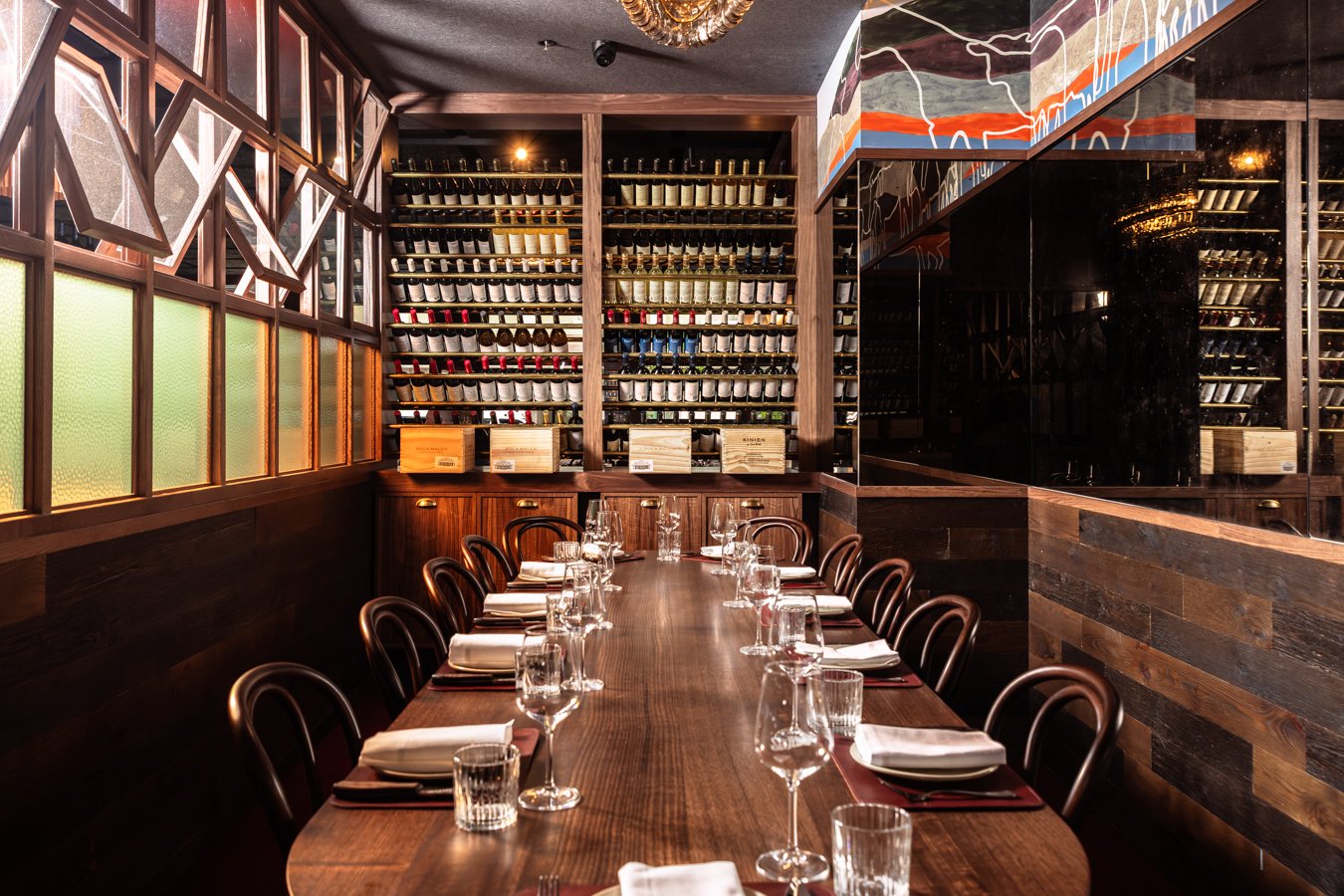 A private wine-tasting room with a long wooden table set with glasses, napkins, and cutlery. The room features a large wall of wine bottles in wooden racks, dark wood paneling, and modern decor with colorful artwork on the ceiling and reflective blac