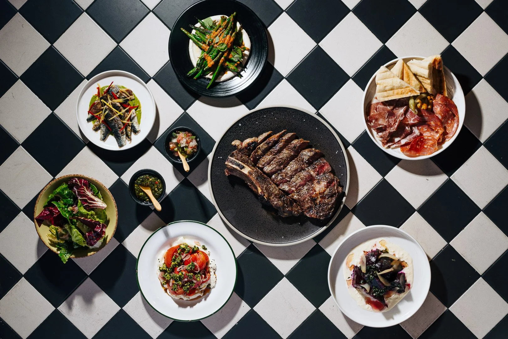 A variety of dishes on a black and white checkered table, including a grilled steak, salads, grilled vegetables, bread, cured meats, and sauces.