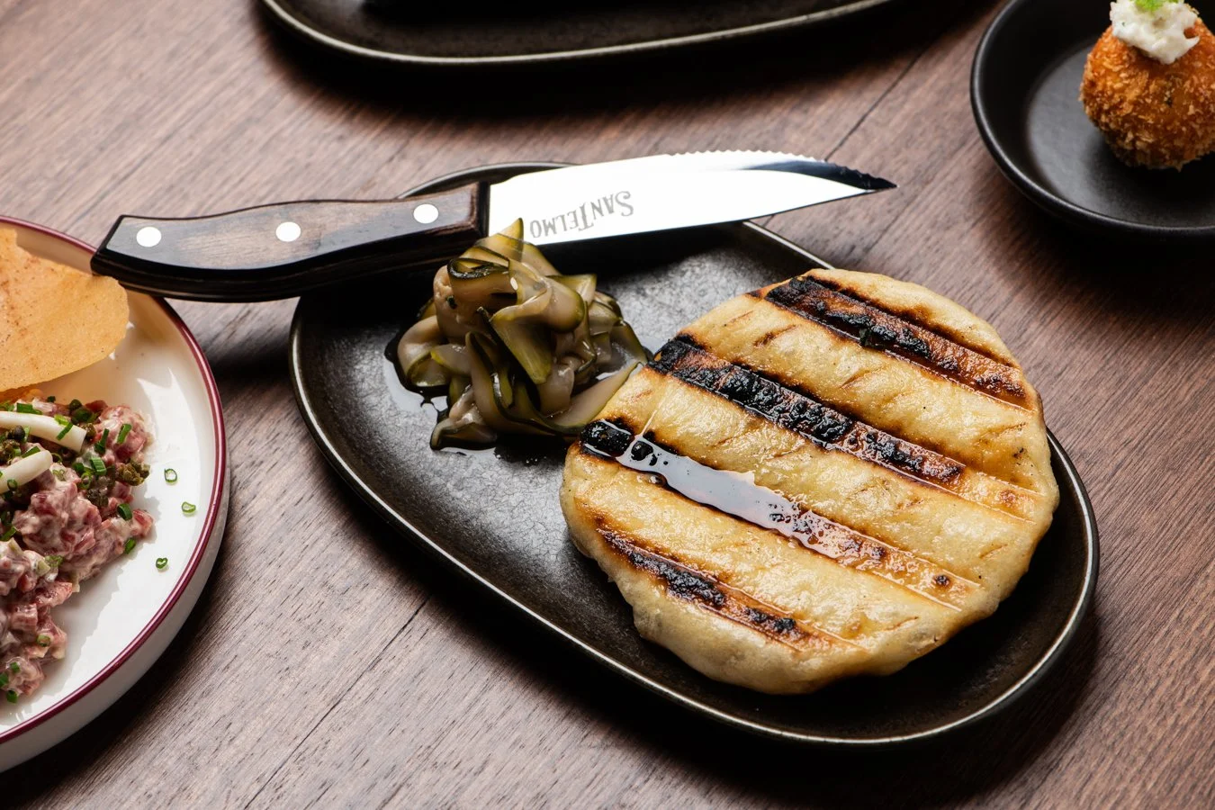 Grilled chicken breast with grill marks served with pickled vegetables on a black plate, with a small side dish of a croquette topped with white sauce on a black dish in the background, and a bowl of salad with herbs on the left.