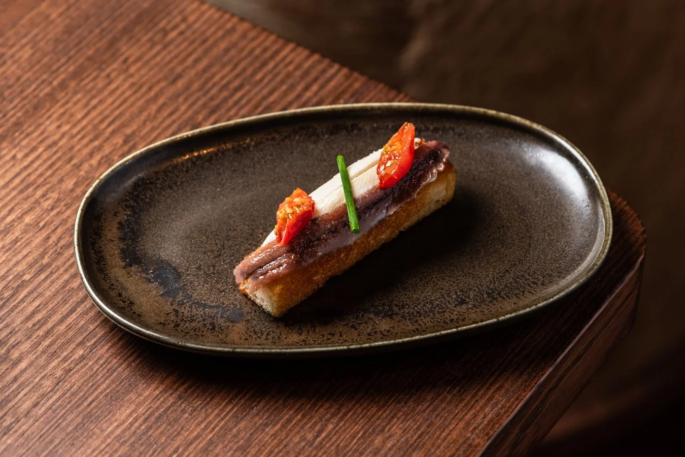A small appetizer with toasted bread, layered with a slice of anchovy, topped with a strip of white cheese, a dot of red chili, and garnished with a chive, on a dark plate.