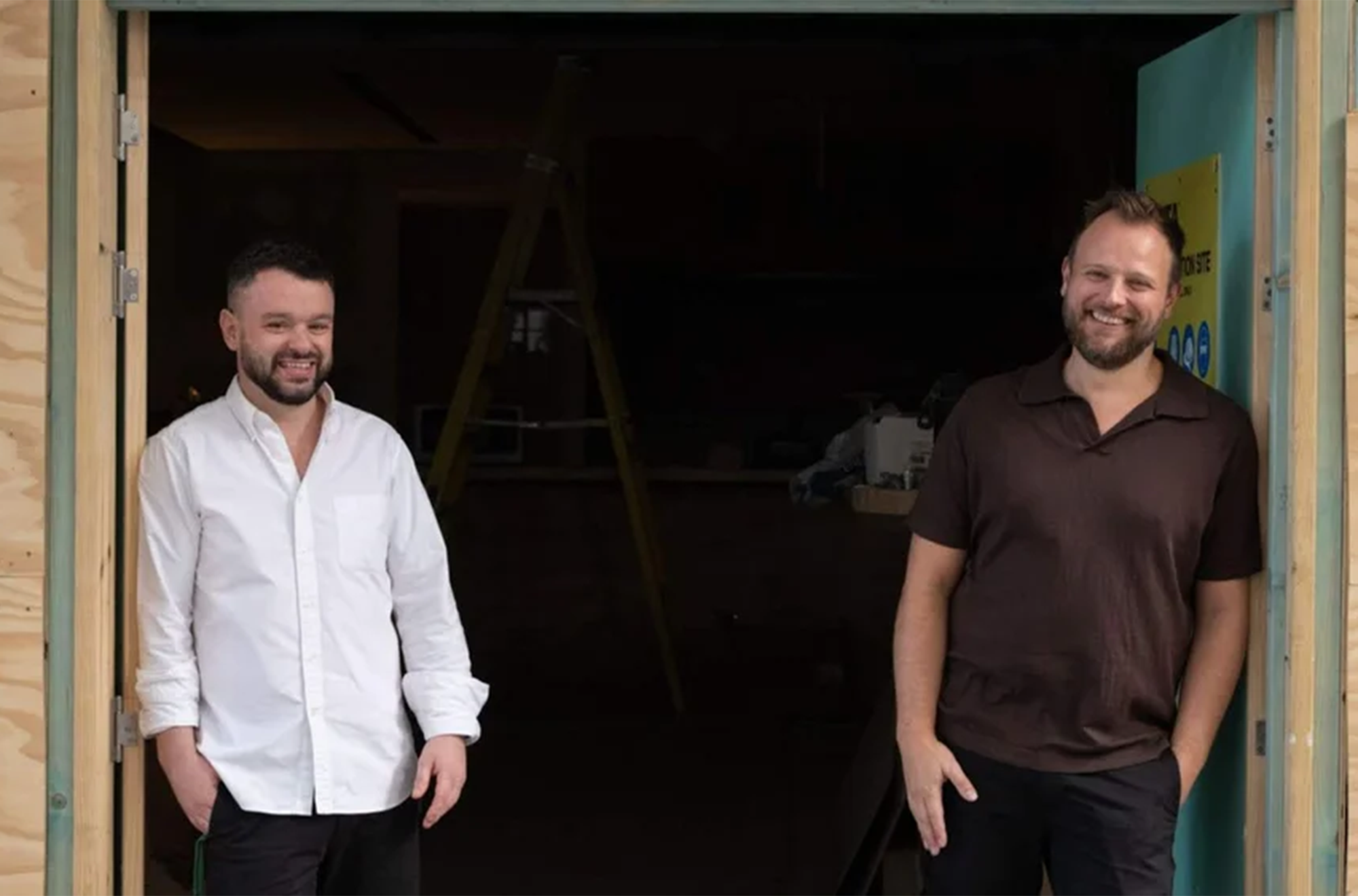Two men stand in a doorway, smiling. The man on the left wears a white shirt, and the man on the right wears a dark Brown shirt. The background shows construction equipment, including a ladder.