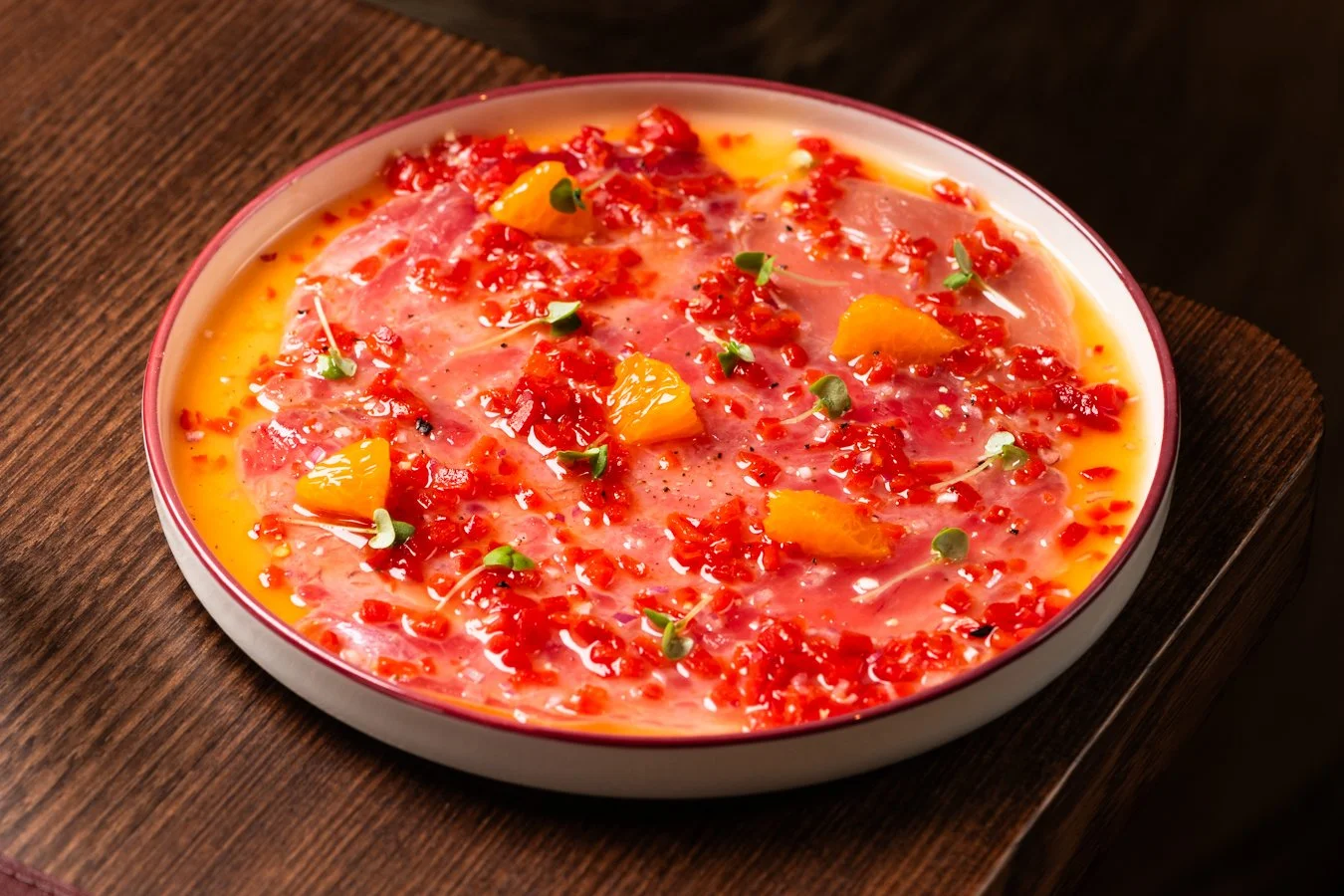 A bowl of raw hamachi fish slices topped with diced red chili, pineapple chunks, microgreens, and red chili flakes.