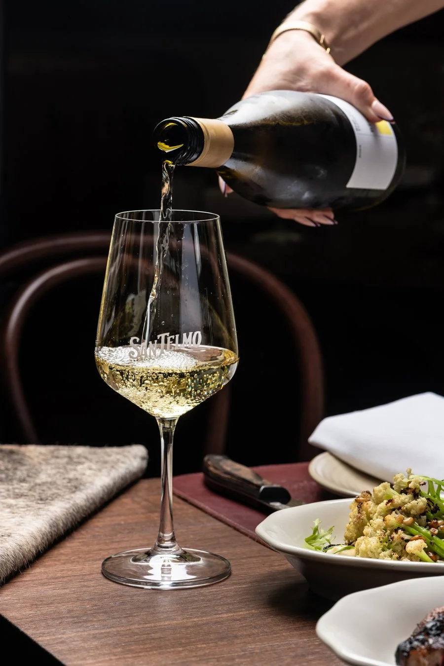 A person pouring champagne into a glass on a dining table with plates of food, including cauliflower, and a plate of cake, in a restaurant setting.