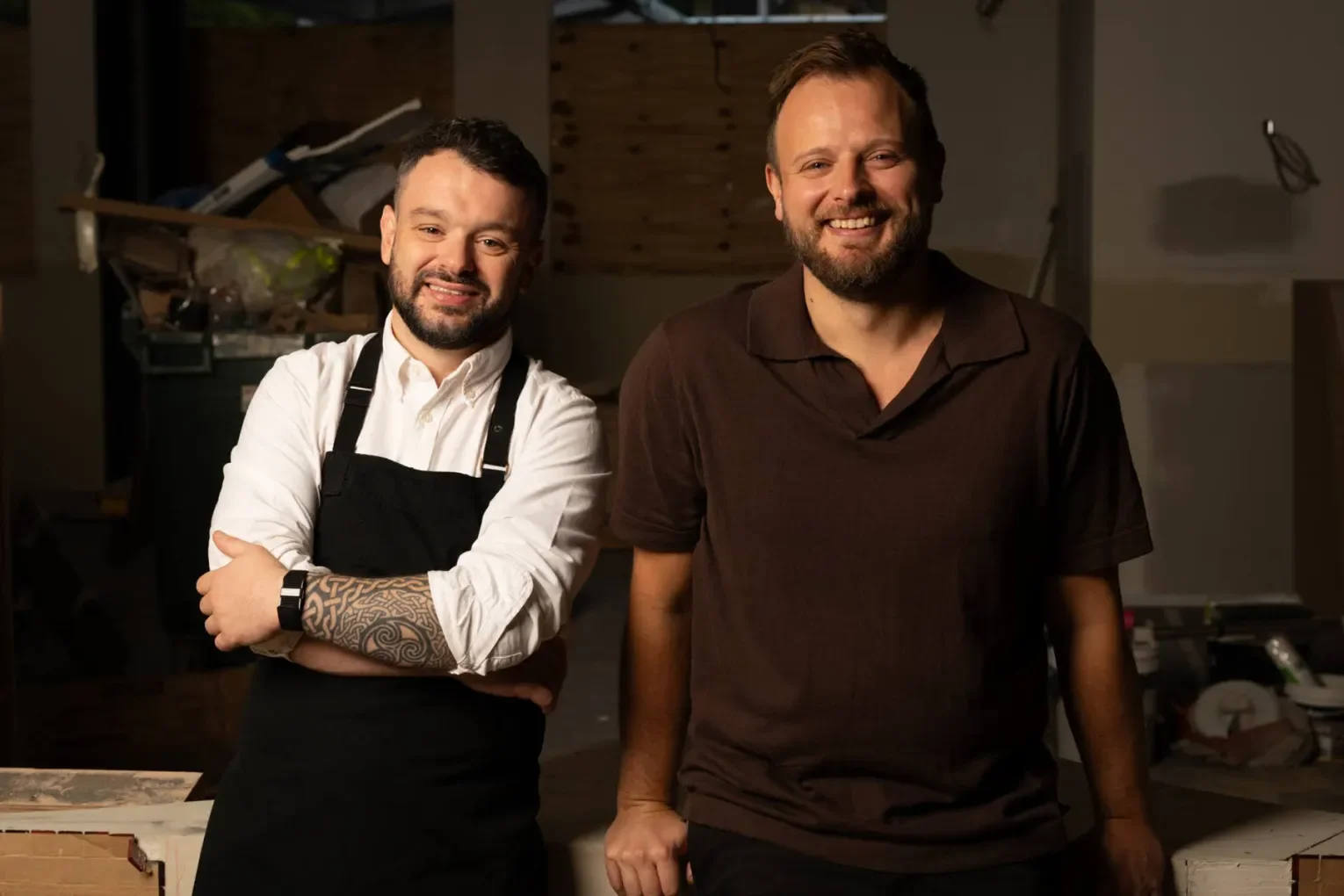Two men smile for the camera in a casual indoor setting. One man wears a white shirt with rolled-up sleeves, a black apron, and has tattoos on his left arm. The other man wears a dark brown polo shirt. The background appears rustic with wooden walls and various items.