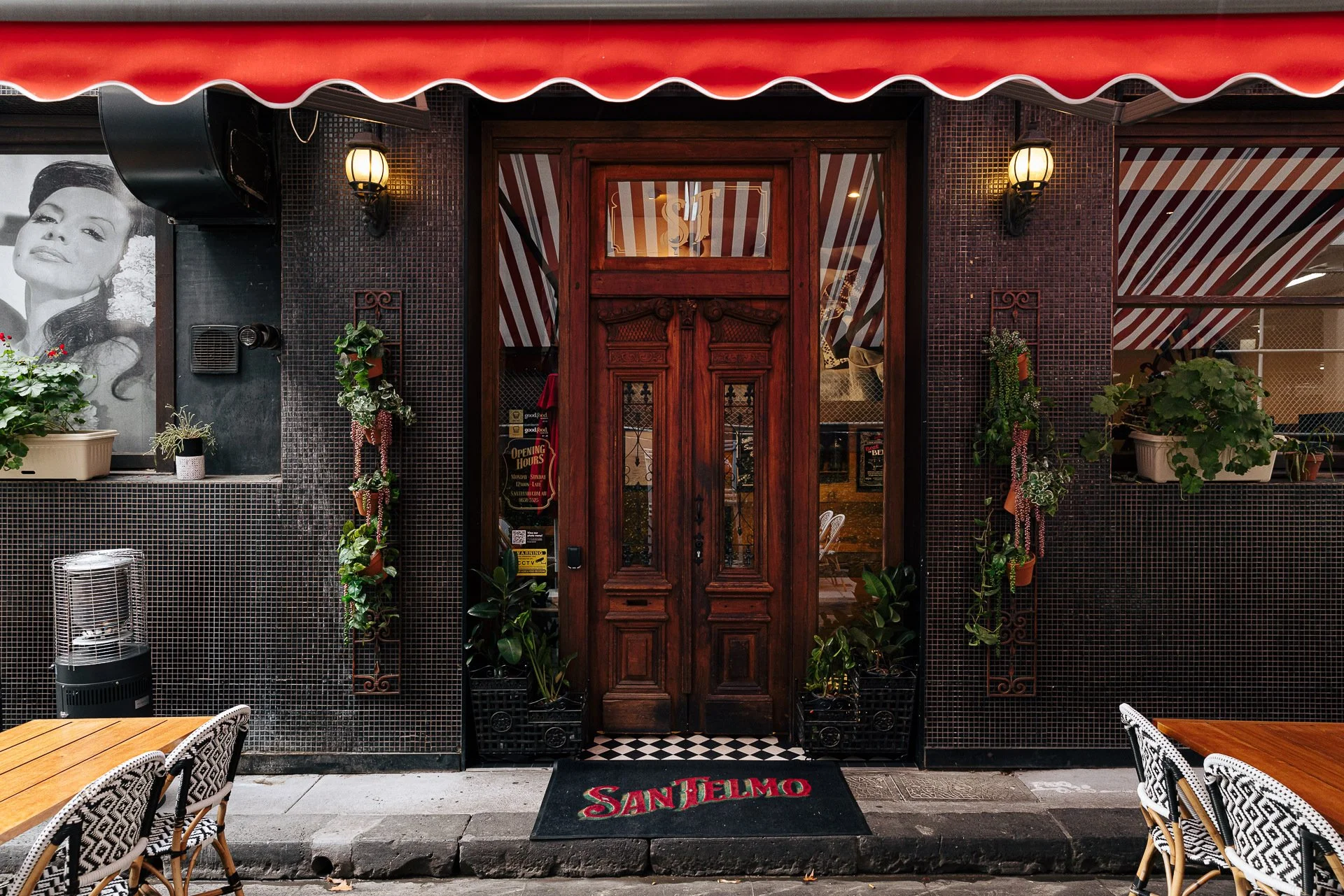 Front entrance of a restaurant with a vintage wooden door, black tile walls, decorative plants, and outdoor seating with patterned chairs and tables. A black and red striped awning is above the door.