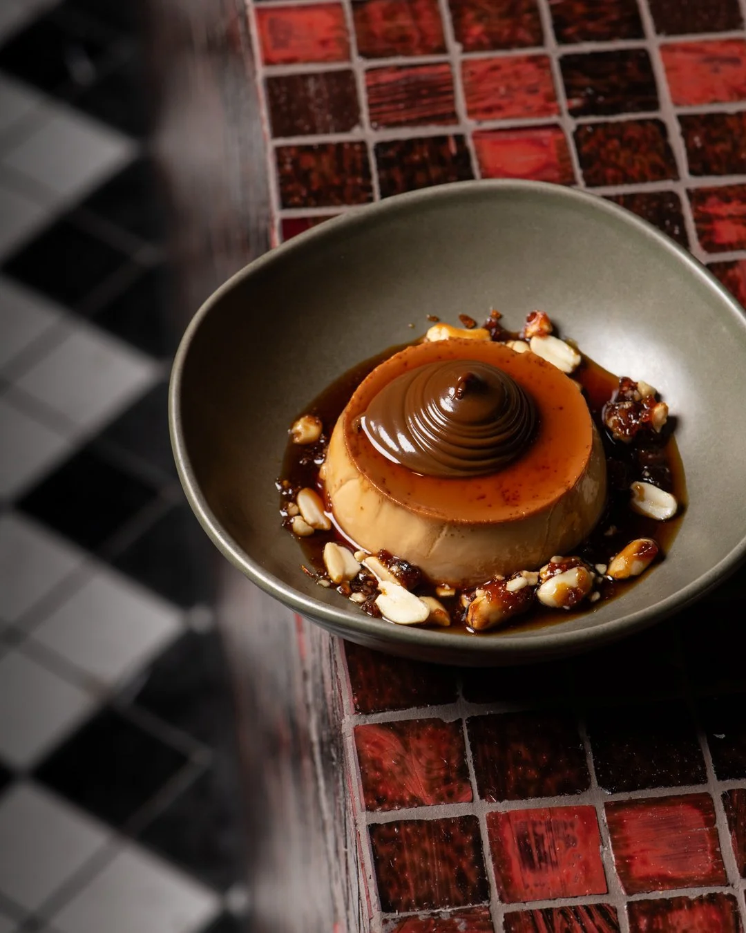 Caramel flan topped with chocolate ganache in a gray bowl, garnished with nuts and caramel sauce, on a red and black tiled surface.
