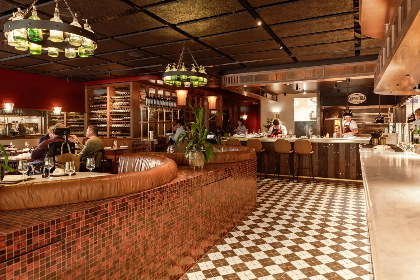 Interior of a cozy restaurant with a bar area, wine racks, dining tables with set glasses, and staff preparing food in the open kitchen.