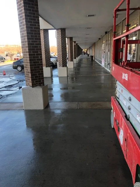 Empty sidewalk outside a retail store with a red lift at the right side. Car parking lot with a few cars is visible to the left.