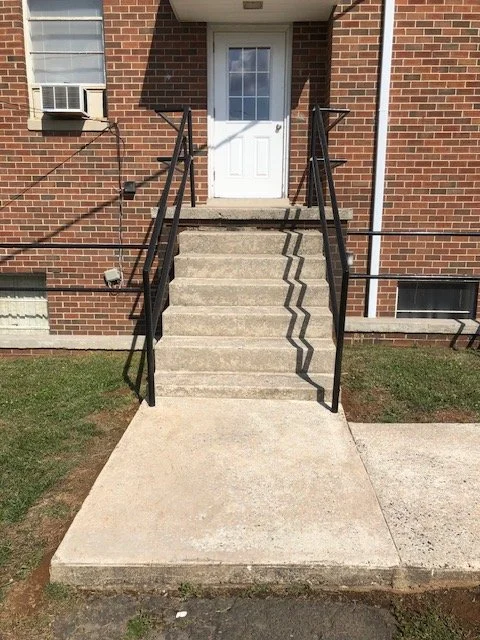 Concrete stairs leading to a white door with black handrails on both sides, brick building exterior, small grassy area and sidewalk in front.