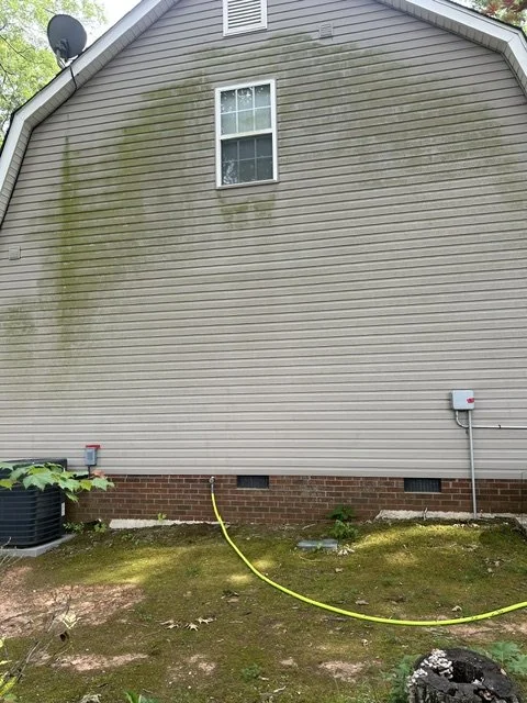 The beige side of a house with a small window, some moss or algae growth on the siding, and a utility box connected by a yellow hose on the ground. There is an HVAC unit to the left and some greenery near the bottom.