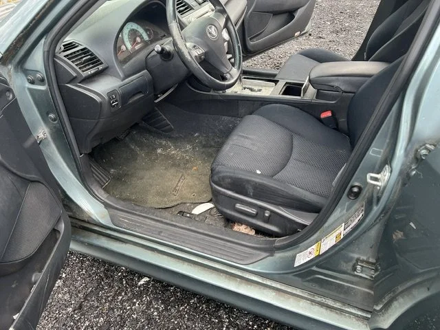 Interior of a car showing the driver's seat, dashboard, steering wheel, and part of the front passenger seat. The vehicle appears dusty and has some dirt on the floor.