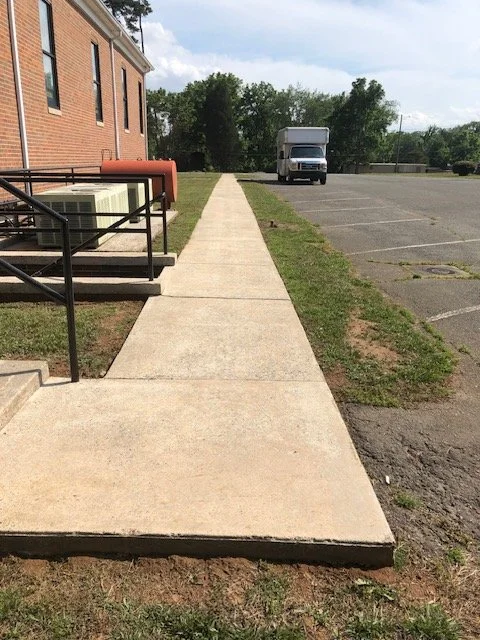 Concrete sidewalk outside a brick building with a metal railing, a red mailbox, and a parking lot with a white delivery truck in the background.