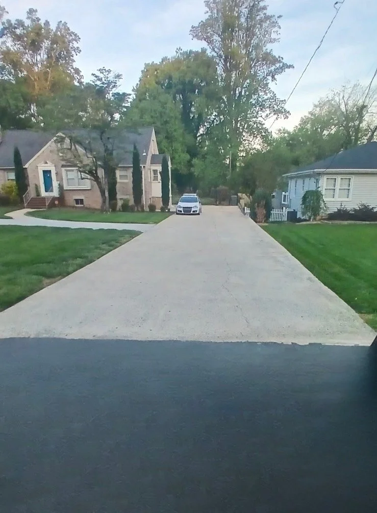 View of a suburban street from inside a vehicle, showing a concrete driveway, green lawns, trees, and houses on both sides, with a car parked at the end of the driveway.