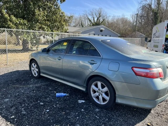 A blue sedan parked on gravel in a lot with a chain-link fence and trees in the background.