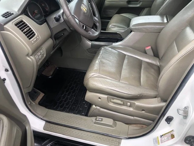 Interior of a car showing the driver's seat, steering wheel, dashboard, and part of the passenger seat. The seats are beige leather, and the floor has black rubber mats.