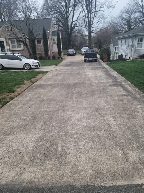 View of a concrete driveway extending from the foreground to the background, flanked by grass on both sides and residential houses with trees in the background.