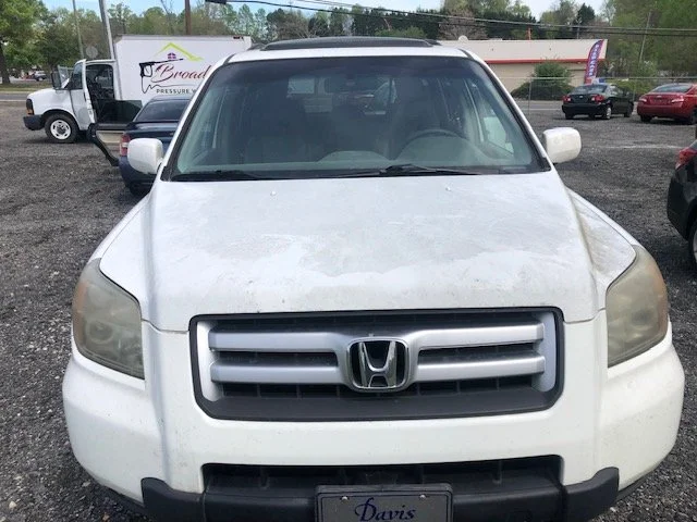 Front view of a white Honda SUV parked on a gravel lot with other vehicles and a dealership sign in the background.