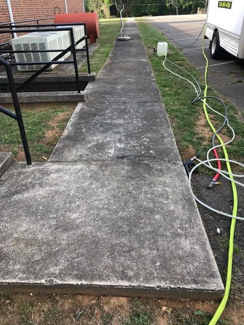 Concrete sidewalk with electrical cords running along the right side, a white utility trailer, and outdoor equipment.