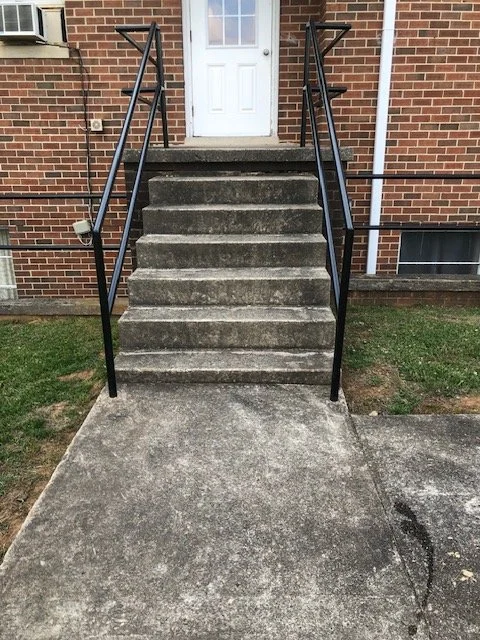 Concrete stairs leading to a white front door with black metal handrails on both sides. The stairs are attached to a brick house, with grass on either side of the sidewalk.